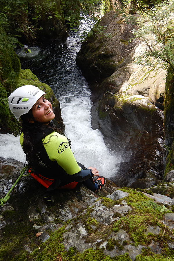 Junge Frau beim Canyoning im Tessin sitzt lächelnd am Felsrand vor einer Schlucht mit Wasserfall – purelements Canyoning Tour